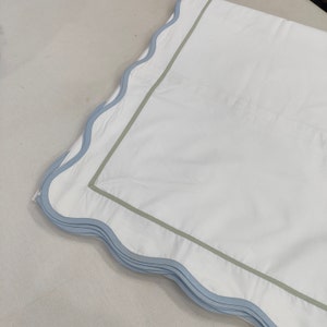 Op de afbeelding: Wit beddengoed met een lichtblauwe geschulpte rand en een saliegroene rand. Het beddengoed heeft een strakke, heldere uitstraling, wat wijst op een focus op comfort en stijl. Het ontwerp is eenvoudig maar elegant.