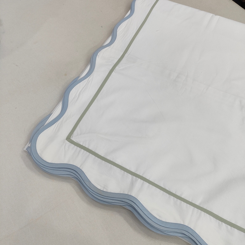 Op de afbeelding: Wit beddengoed met een lichtblauwe geschulpte rand en een saliegroene rand. Het beddengoed heeft een strakke, heldere uitstraling, wat wijst op een focus op comfort en stijl. Het ontwerp is eenvoudig maar elegant.