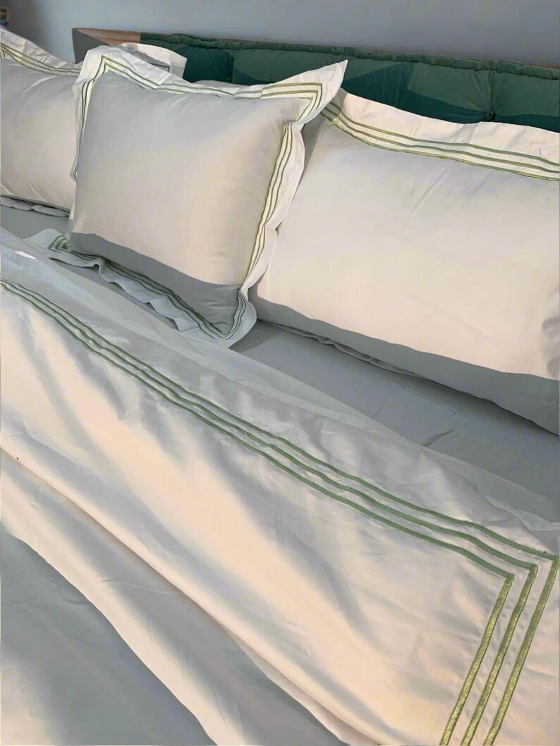 May include: White bed linen with green trim. The linen is folded and arranged on a bed with a green headboard.