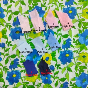Op de afbeelding: Negen stoffen stalen in verschillende roze en blauwe tinten worden getoond op een bloemmotief achtergrond. De stalen zijn gelabeld met kleurnamen zoals "Baby Pink", "Lichtblauw" en "Marineblauw". De achtergrond heeft blauwe bloemen en groene bladeren.