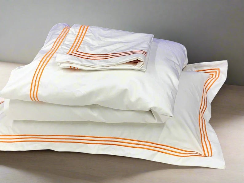 May include: A stack of white pillowcases with orange trim. The pillowcases are folded and arranged on a light wood surface.