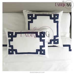 May include: White decorative pillows with a navy blue geometric border. The pillows are rectangular and square, placed on a white bedspread. The design is a modern take on a classic style, suitable for bedroom decor.