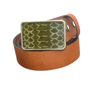 May include: A brown leather belt with a rectangular buckle. The buckle has a gold-colored frame and a green, scale-patterned center. The belt is made of a light brown leather and has a classic design.