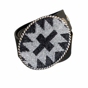 May include: A black leather belt with a silver oval buckle featuring a gray and black geometric pattern.