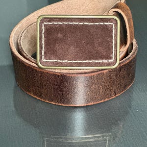May include: A brown leather belt with a brass buckle. The buckle has a brown suede insert with white stitching.