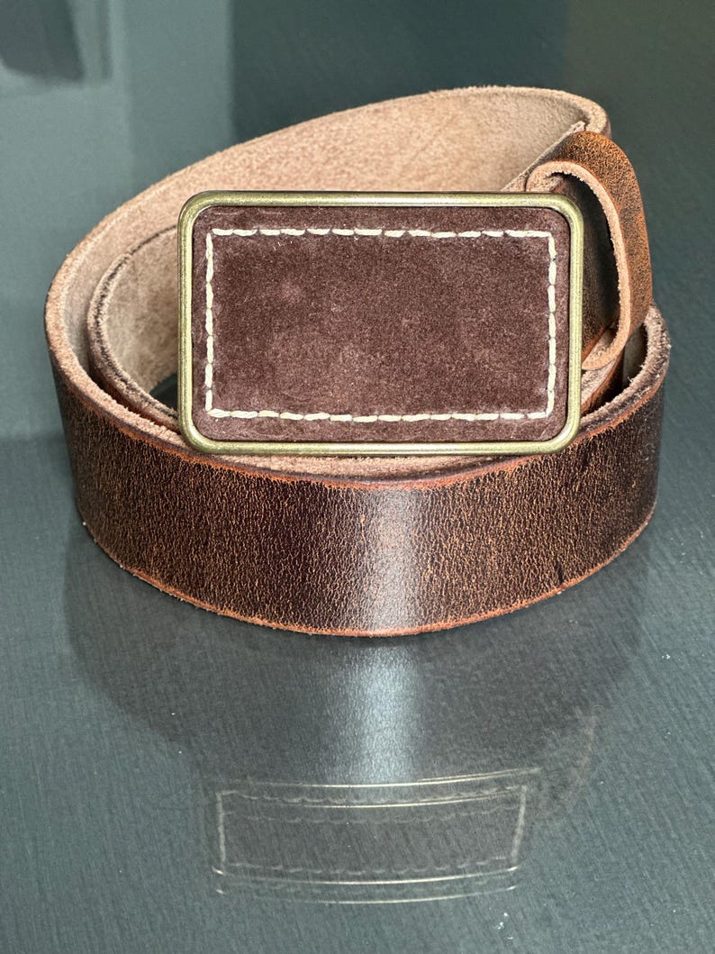 May include: A brown leather belt with a brass buckle. The buckle has a brown suede insert with white stitching.
