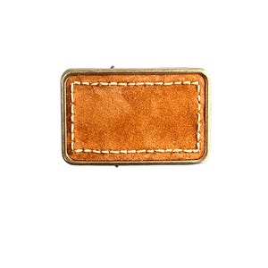 May include: A rectangular brown leather patch with a stitched border. The patch is surrounded by a metal frame.