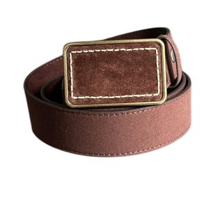May include: A brown suede belt with a brass buckle. The buckle has a suede insert with a stitched border.