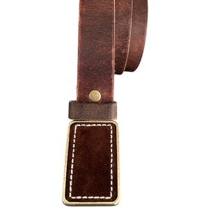 May include: A brown leather belt with a brass buckle. The buckle has a brown leather insert with white stitching around the edges.