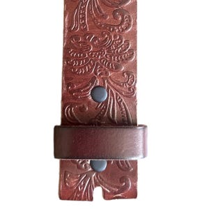 May include: A brown leather belt with an embossed floral pattern. The belt has a metal buckle and two black rivets.