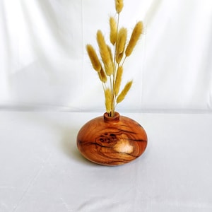 May include: A wooden vase with a round, bulbous shape and a natural wood finish. The vase has a unique, knotty grain pattern and is filled with dried, light brown pampas grass.