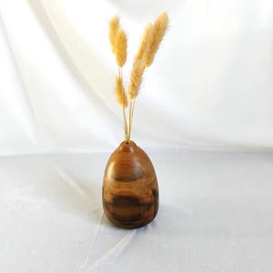 May include: A wooden vase with a rounded shape, holding several dried, fluffy, yellow-tan stems. The vase has a rich, brown wood grain pattern. The background is a white fabric.