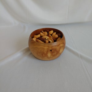 May include: A small, round wooden bowl filled with a mix of roasted nuts. The bowl is made of light brown wood and has a smooth finish. The nuts include cashews and other varieties, with a mix of light and dark brown colors.