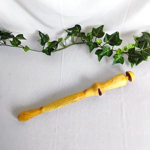 May include: A handcrafted wooden rolling pin with a natural, light yellow finish. The rolling pin features a unique, organic shape with a handle and a decorative knob. The background is a white surface with green ivy.
