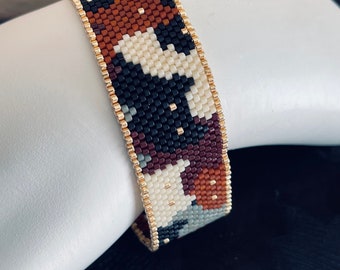 May include: A beaded bracelet featuring a geometric pattern in shades of brown, black, white, and grey. The bracelet is crafted with small, square beads and has a gold-coloured edging.