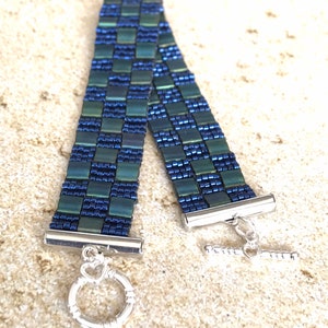 May include: A green and blue beaded bracelet with a silver toggle clasp. The bracelet is made of small, square beads that are arranged in a checkered pattern.