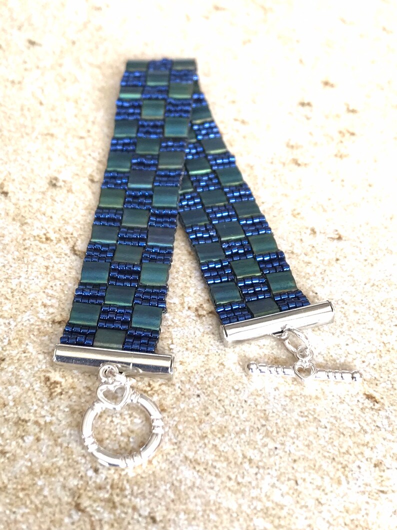 May include: A green and blue beaded bracelet with a silver toggle clasp. The bracelet is made of small, square beads that are arranged in a checkered pattern.