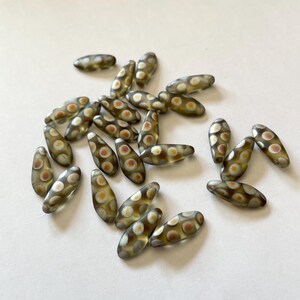 May include: A collection of oval-shaped glass beads. The beads are transparent with a light brown hue and feature a pattern of white and orange dots. The beads are scattered on a white surface.
