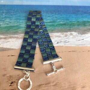 May include: A blue and green beaded bracelet with a silver toggle clasp. The bracelet is made of small, square beads that are arranged in a checkered pattern.