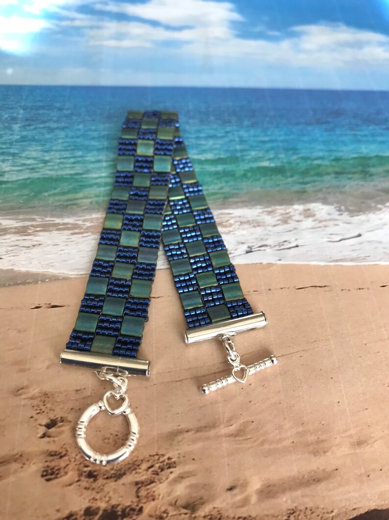 May include: A blue and green beaded bracelet with a silver toggle clasp. The bracelet is made of small, square beads that are arranged in a checkered pattern.