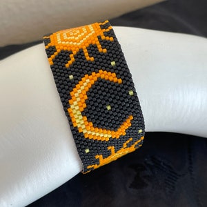 May include: A beaded bracelet featuring a celestial design. The bracelet is primarily black, with orange and yellow accents forming a crescent moon, stars, and sun pattern. The bracelet is made of small, evenly spaced beads.