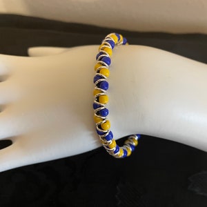 May include: A yellow and blue beaded bracelet with a silver wire coil. The bracelet is on a white mannequin hand.