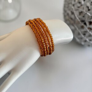 May include: A multi-strand wrap bracelet composed of small, translucent amber-colored glass seed beads. The bracelet features several circular rows of uniform, shiny beads that create a layered appearance around the wrist area of the display form.