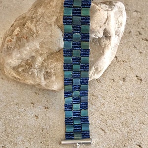 May include: A blue and green beaded bracelet with a silver clasp. The bracelet is made of small, square beads that are arranged in a checkered pattern.