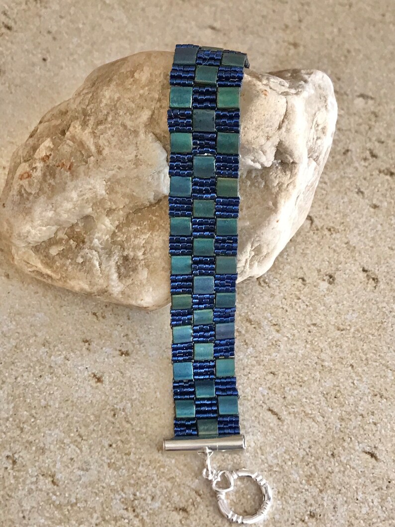 May include: A blue and green beaded bracelet with a silver clasp. The bracelet is made of small, square beads that are arranged in a checkered pattern.