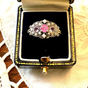 Peut inclure: Une bague vintage avec une pierre rose centrale, flanquée de deux petites pierres roses et entourée de petites perles. La bague est sertie dans un métal argenté et présentée dans un écrin noir avec un fermoir doré.