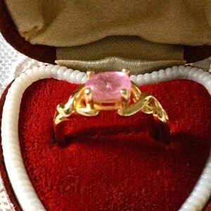 May include: A gold ring featuring a large, oval pink gemstone, set within an ornate filigree design. The ring is presented in a red velvet heart-shaped box with white trim, creating a romantic display.