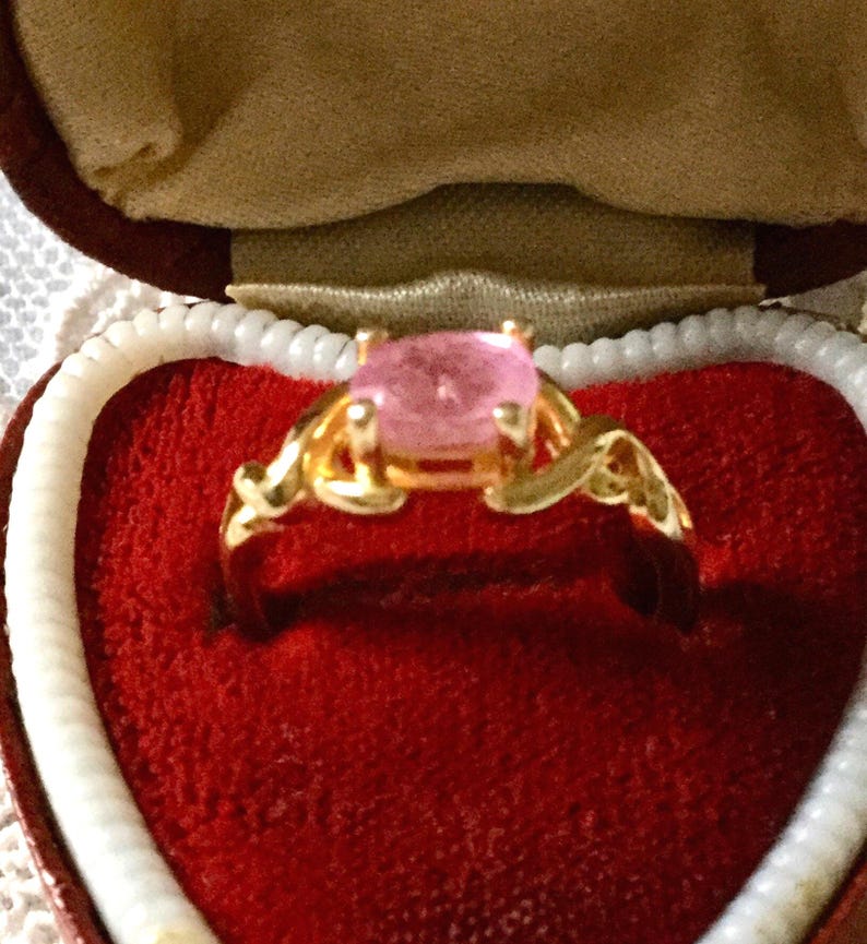 May include: A gold ring featuring a large, oval pink gemstone, set within an ornate filigree design. The ring is presented in a red velvet heart-shaped box with white trim, creating a romantic display.