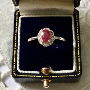 Puede incluir: Un anillo de plata con una piedra ovalada roja rodeada de piedras blancas más pequeñas. El anillo está en una caja de anillo de terciopelo azul.