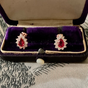 Peut inclure: Boucles d'oreilles en or rose avec une pierre rouge en forme de goutte d'eau, entourée de petites pierres claires. Les boucles d'oreilles sont présentées dans un écrin en velours violet ouvert.