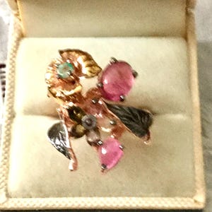 Peut inclure: Une bague ornée d'un motif floral, en métal doré, ornée de pierres roses, d'accents verts et noirs. La bague est présentée dans un écrin beige, mettant en valeur son design complexe et son artisanat.