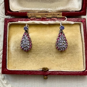 Peut inclure: Une paire de boucles d'oreilles en forme de goutte avec un sertissage pavé de petites pierres violet clair et rose foncé. Chaque boucle est surmontée d'une pierre violet clair plus grande et d'une attache à levier argentée.