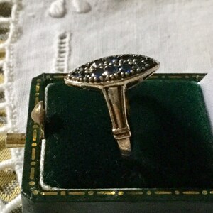 May include: A gold-toned ring with a marquise-shaped top, set with numerous small, dark blue gemstones. The ring is displayed in a green velvet jewelry box, showcasing its elegant design and craftsmanship.