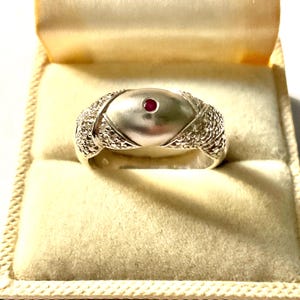 Peut inclure: Une bague en argent avec un design central ovale et une petite pierre précieuse rouge ronde. La bague présente des détails texturés sur les côtés et est présentée dans un écrin crème.