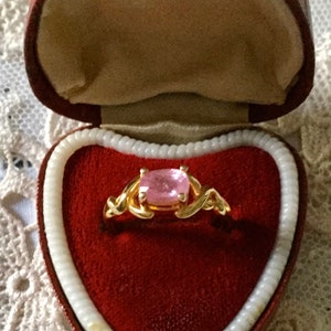May include: A gold ring with a square, pale pink gemstone, displayed in a heart-shaped red velvet box. The ring's band has an intricate, swirling design. The box is trimmed with cream-coloured edging and rests on a lace surface.