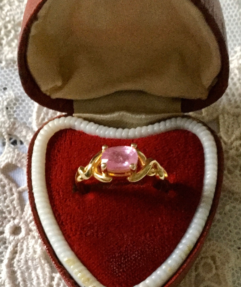 May include: A gold ring with a square, pale pink gemstone, displayed in a heart-shaped red velvet box. The ring's band has an intricate, swirling design. The box is trimmed with cream-coloured edging and rests on a lace surface.