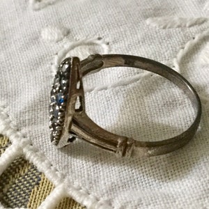 May include: A silver ring with a cluster of small, round, clear and blue gemstones. The ring has a decorative, openwork design on the sides and is set against a white, textured background.