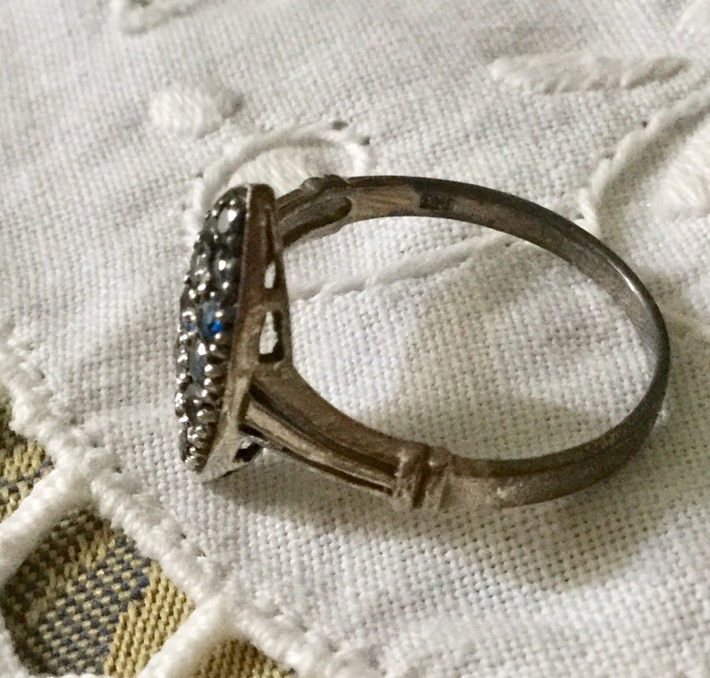 May include: A silver ring with a cluster of small, round, clear and blue gemstones. The ring has a decorative, openwork design on the sides and is set against a white, textured background.
