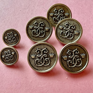 Peut inclure: Ensemble de boutons anciens de couleur laiton. Les boutons ronds présentent un motif décoratif avec un monogramme stylisé et de petites couronnes. Différentes tailles de boutons, sur fond rose.