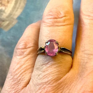 Peut inclure: Une bague en argent ornée d'une grosse pierre rose de forme ovale. Le corps de la bague présente un design simple avec un accent sombre de chaque côté de la pierre précieuse. La bague est portée sur un doigt.