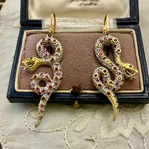 Peut inclure: Une paire de boucles d'oreilles serpent en or et argent, ornées de pierres précieuses rouges et vertes. Les boucles d'oreilles sont présentées dans un écrin bleu avec un intérieur rose. Les boucles d'oreilles serpent sont détaillées avec des pierres de couleur rubis et des yeux verts.