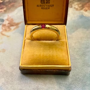 Peut inclure: Une bague en or avec une seule pierre rouge, présentée dans un écrin vintage avec un intérieur en velours jaune. La boîte porte du texte et un blason sur le couvercle. La bague est au centre.