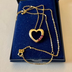 Könnte beinhalten: Eine goldene Halskette mit einem herzförmigen Anhänger, der mit drei kleinen roten Edelsteinen besetzt ist. Die Halskette wird in einer blauen Samtbox präsentiert.