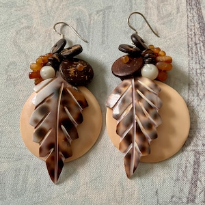 Peut inclure: Une paire de boucles d'oreilles artisanales avec un motif de feuille. Les boucles d'oreilles ont une base circulaire beige avec un accent de feuille marron et crème. Des perles en bois marron et des crochets d'oreilles argentés complètent le design.