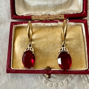 Peut inclure: Paire de boucles d'oreilles dorées avec de grandes pierres rouges ovales et facettées. Les boucles d'oreilles sont présentées dans un écrin rouge avec un intérieur crème.