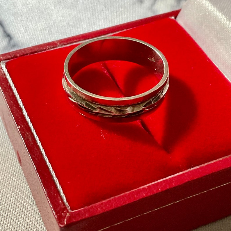May include: A silver ring with a textured band, presented in a red jewelry box. The ring's band features a repeating pattern. The ring is displayed against a red velvet interior.